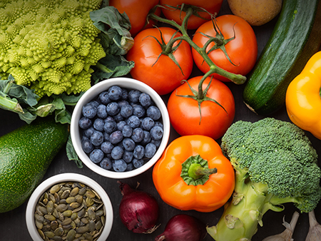 Tomaten, Brokkoli, Paprika, Zucchini, Blaubeeren, Nüsse und anderes Obst und Gemüse liegen dicht gedrängt auf einem Tisch.